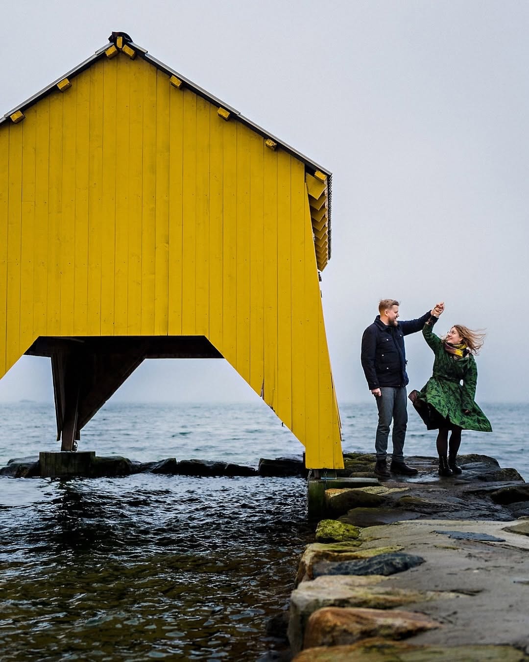 A couple beside a bright yellow boathouse on an overcast day