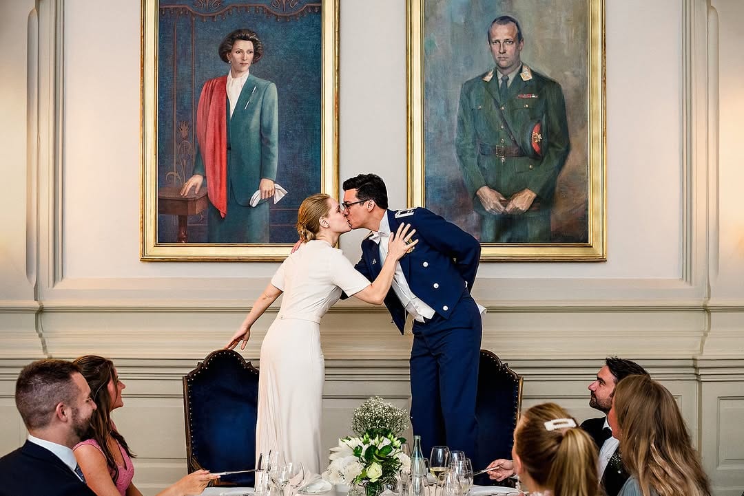 A couple kissing beside a dining table while guests sit nearby