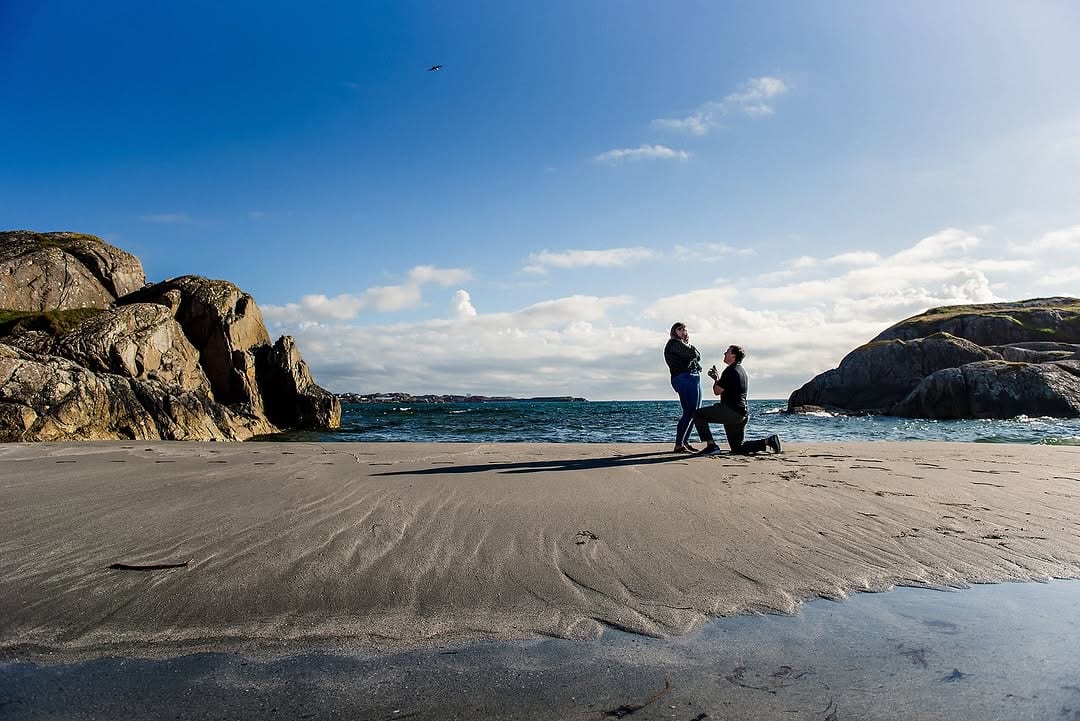 A couple on a sandy beach beside rocky outcrops during a proposal