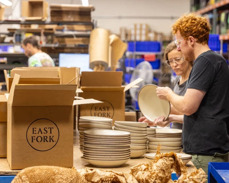 People handling ceramic plates beside stacks of dishes and open cardboard boxes.