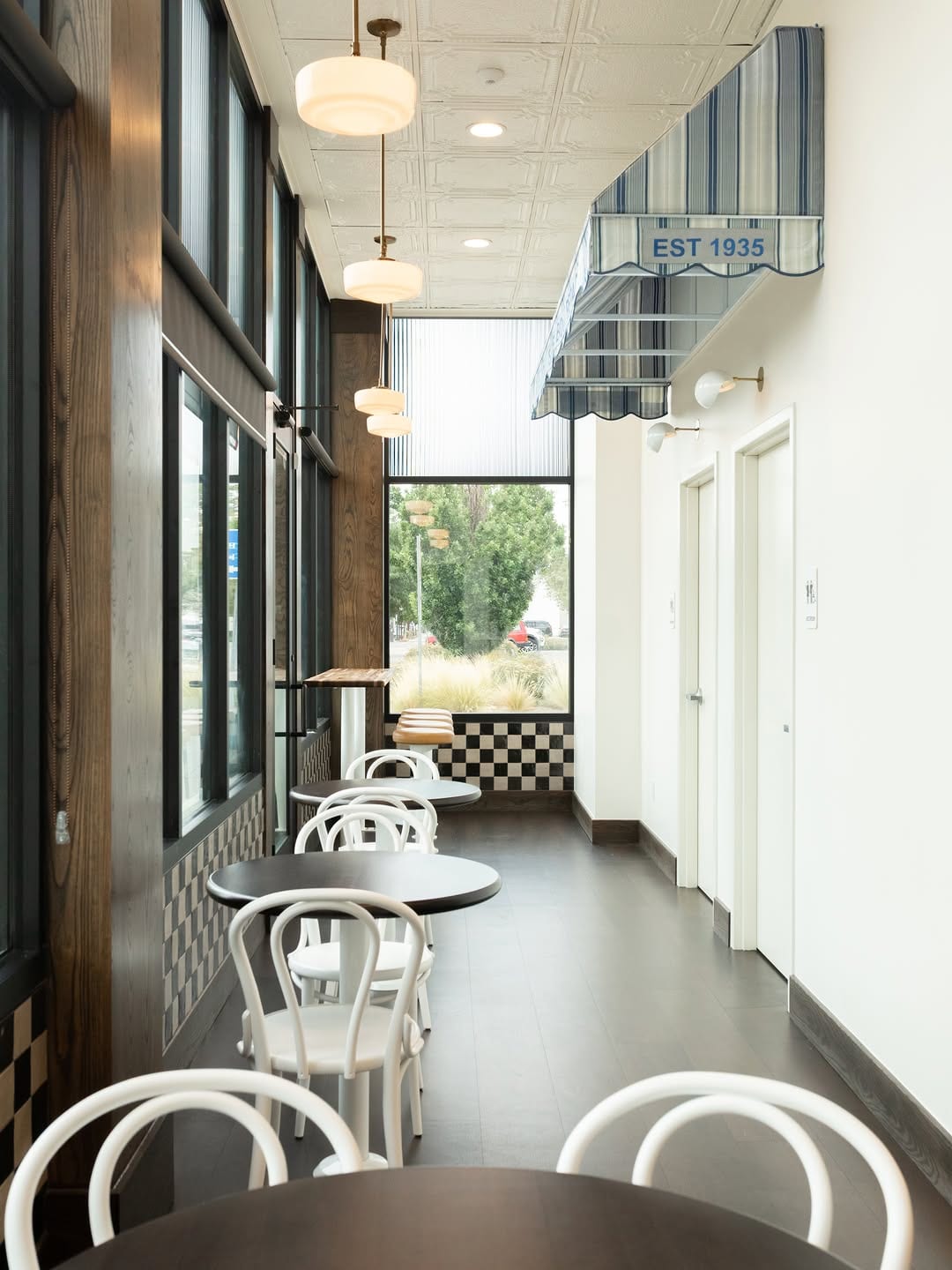 Narrow café seating area with white chairs, round tables, pendant lights and tall windows.