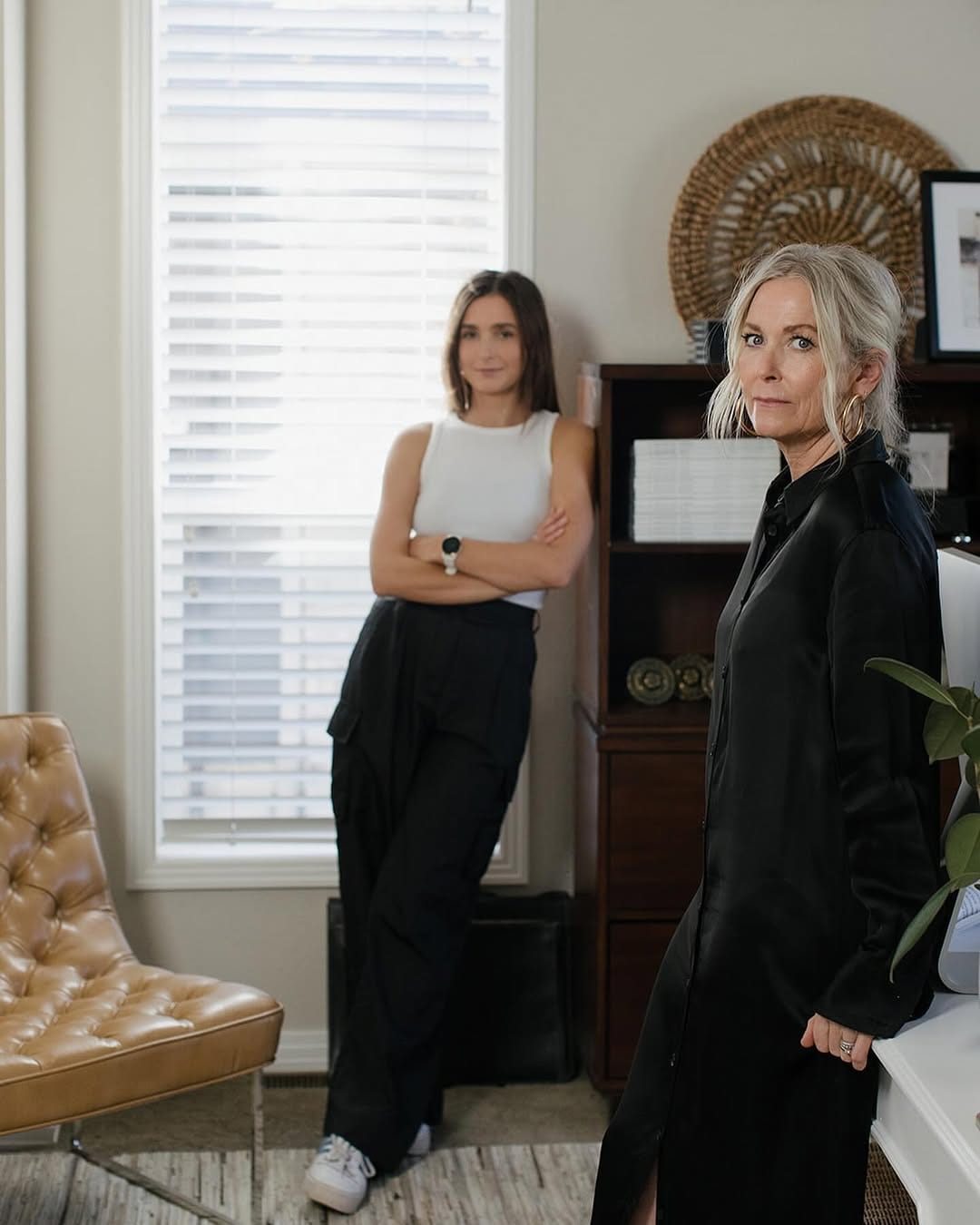 Two women standing indoors near a window with shelving, framed art and houseplants.