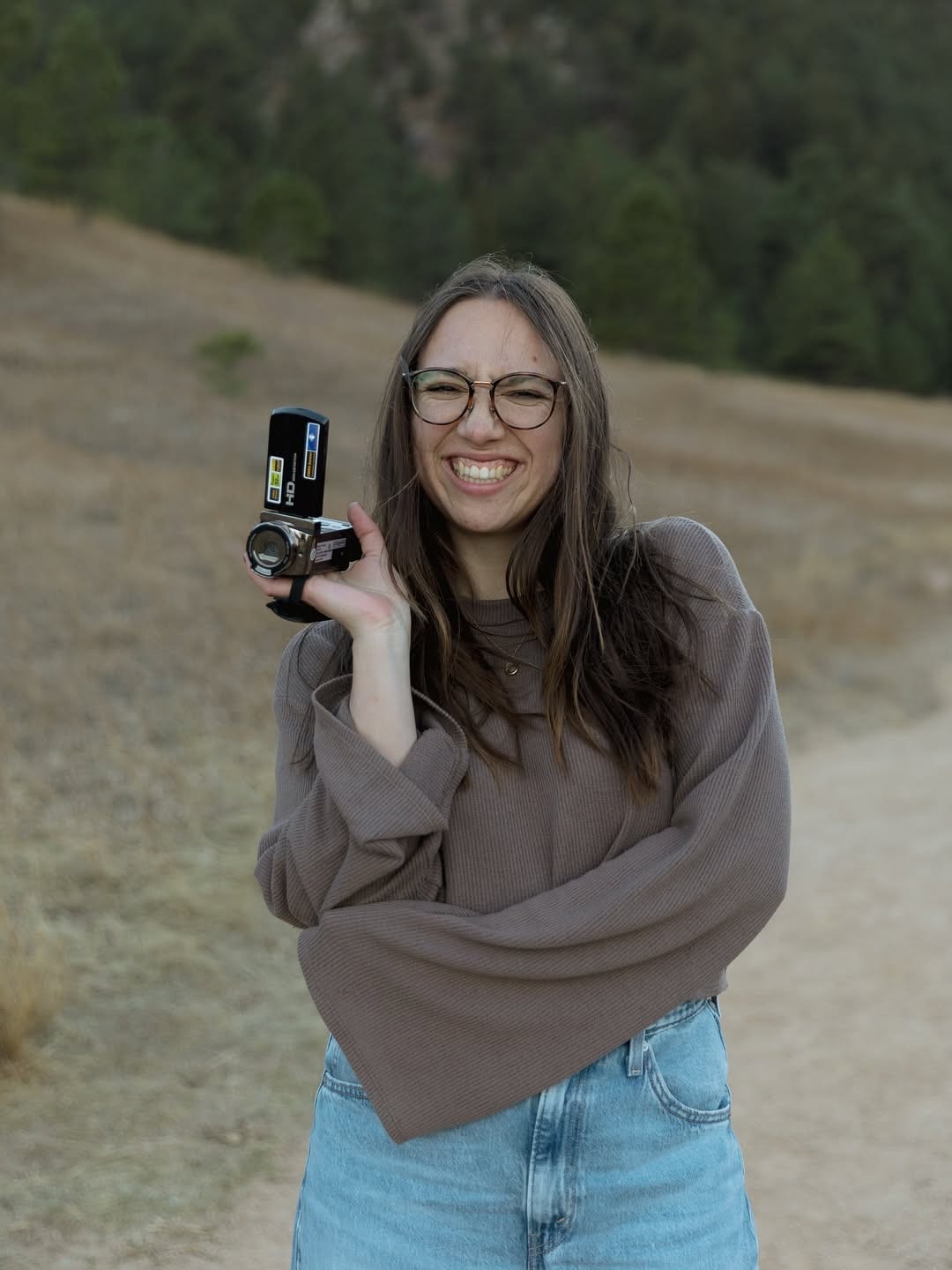 Smiling Kira holding a small camera on a wooded hillside